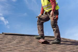 Unrecognizable workman standing on tile roof of new home under construction against blue sky with helmet in hands