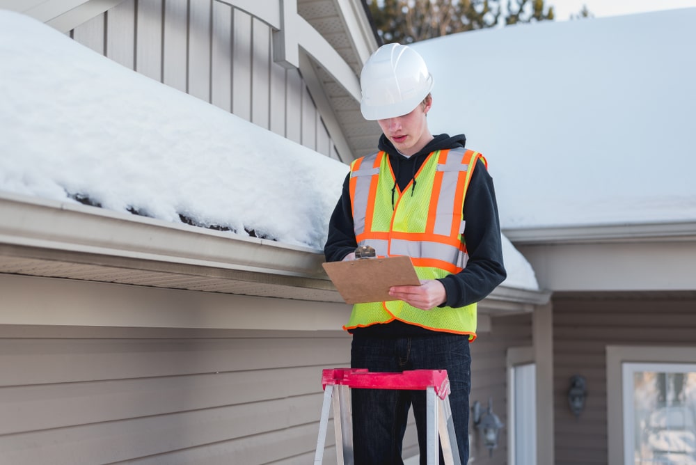Home inspector on a ladder while writing on a clipboard during winter.