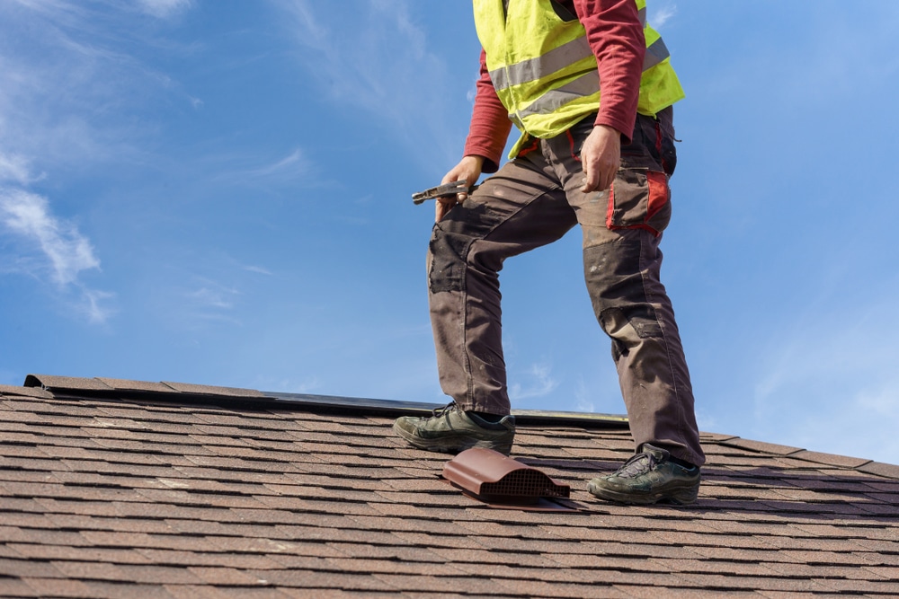 Unrecognizable workman standing on tile roof of new home under construction against blue sky with helmet in hands